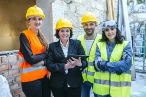Coordinadora de Actividades Empresariales en traje formal con tablet revisando medidas de seguridad junto a profesionales con EPIs en obra de construcción