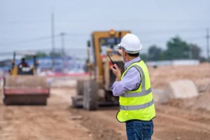 Trabajador con chaleco reflectante y casco actuando como recurso preventivo en una obra, supervisando maquinaria pesada y garantizando la seguridad laboral.