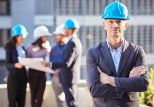 Profesional con casco azul de seguridad y traje ejerciendo como coordinador de actividades empresariales, mostrando liderazgo mientras supervisa un equipo técnico que revisa documentación en una obra