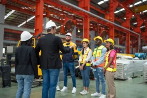 Profesionales con cascos de seguridad participando en una reunión de coordinación empresarial en planta industrial, con líderes explicando procedimientos a trabajadores con chalecos reflectantes