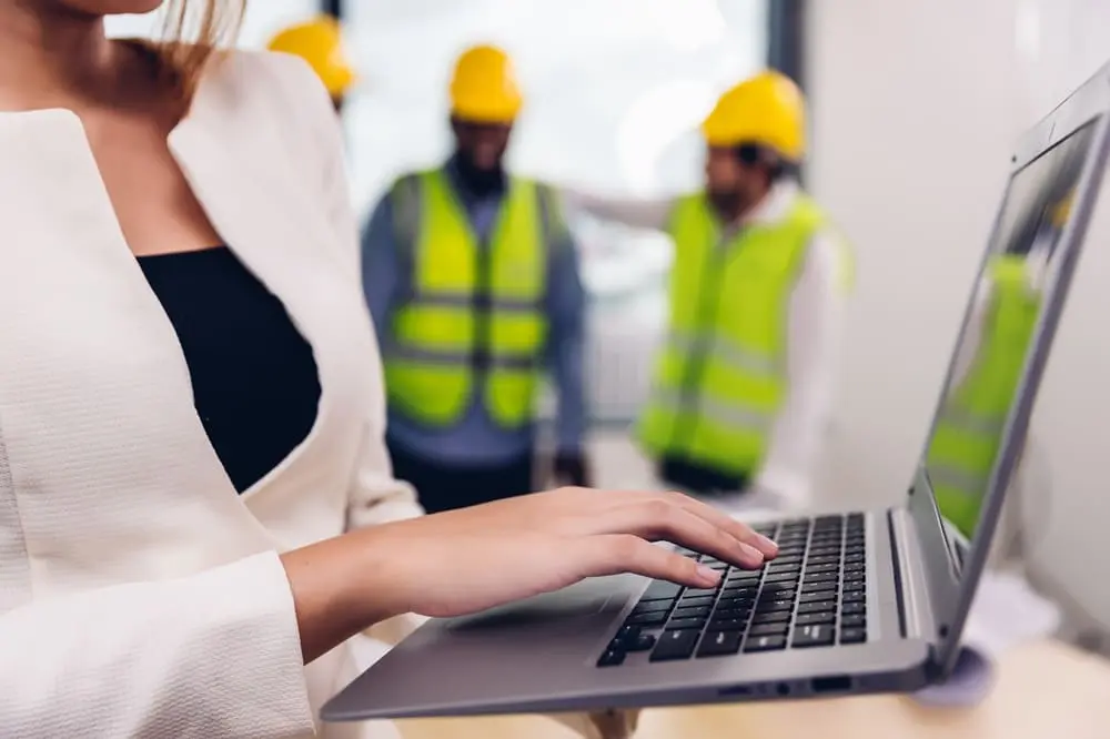 Técnica de prevención de riesgos laborales gestionando la validación automática de documentos CAE en portátil con trabajadores de construcción de fondo