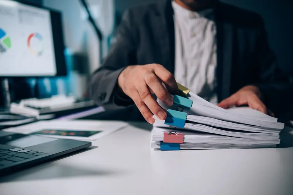 Profesional gestionando documentación CAE manualmente con pilas de documentos en papel - Gestión documental tradicional