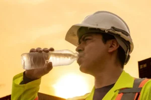 Trabajador de la construcción con casco y chaleco reflectante bebiendo agua de una botella para protegerse del golpe de calor en obra