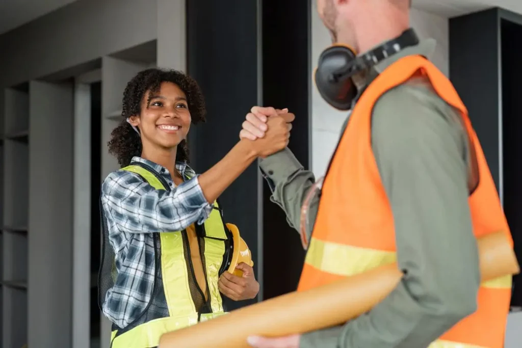 Trabajador y trabajadora de una contrata con equipos de protección se dan la mano en obra, representando el cumplimiento de contratistas antes de la vuelta a la actividad