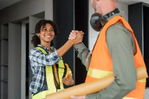Trabajador y trabajadora de una contrata con equipos de protección se dan la mano en obra, representando el cumplimiento de contratistas antes de la vuelta a la actividad
