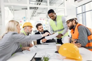 Team multietnico di professionisti edili con caschi di sicurezza e gilet ad alta visibilità che discute piani di cantiere, in conformità con le nuove normative DL 159/2025