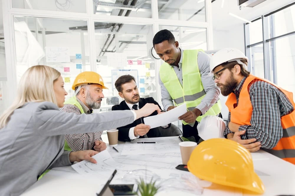 Team multietnico di professionisti edili con caschi di sicurezza e gilet ad alta visibilità che discute piani di cantiere, in conformità con le nuove normative DL 159/2025