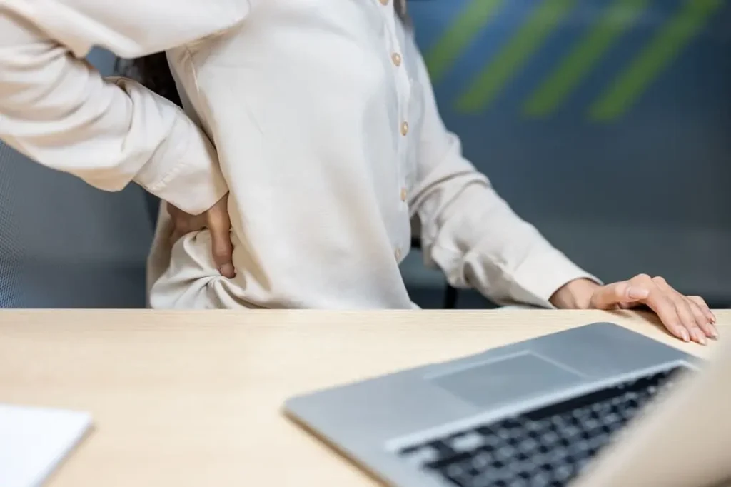 Persona en camisa blanca sentada frente a laptop experimentando dolor de espalda, ilustrando problemas comunes de ergonomía laboral en oficina.