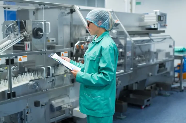 Profissional de controlo de qualidade farmacêutica a realizar auditoria em sala limpa com equipamento de produção conforme Boas Práticas de Fabrico GMP