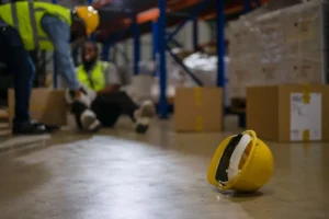 Acidente de trabalho em armazém — trabalhador caído no chão com capacete amarelo em primeiro plano