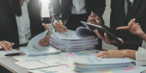 Equipa de profissionais a rever pilhas de documentos em papel sobre uma mesa de trabalho, representando a gestão manual de autorizações de trabalho